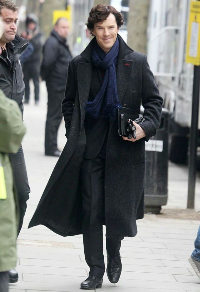 a man walking down the street with a camera in his hand while wearing a long coat and scarf