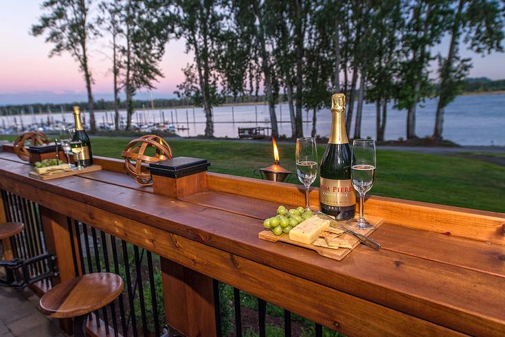 two bottles of wine and some cheese on a wooden table outside with water in the background