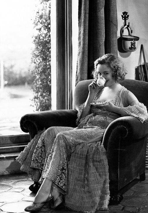 an old photo of a woman sitting in a chair talking on the phone and drinking from a cup