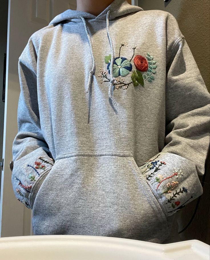 a person wearing a grey hoodie with embroidered flowers on the front and sleeves, standing next to a sink