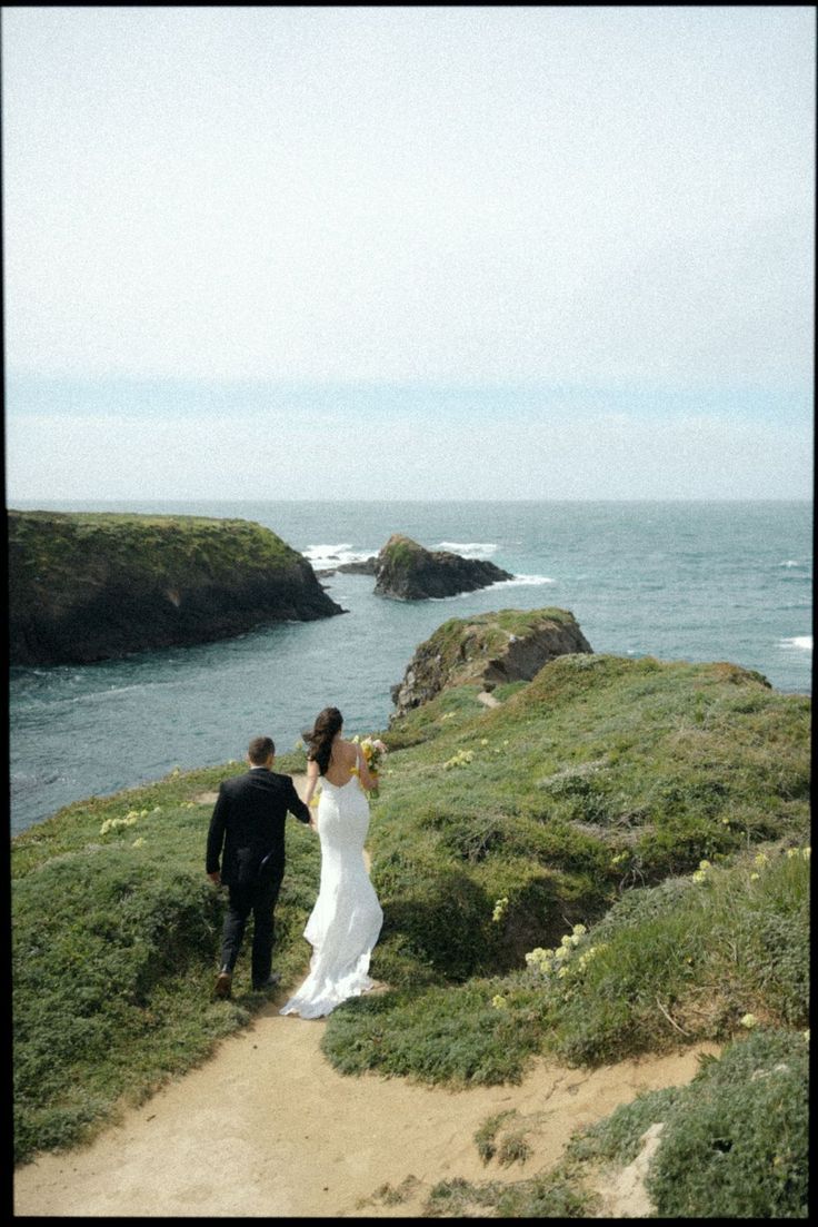 a bride and groom walking down a path towards the ocean with cliffs in the background