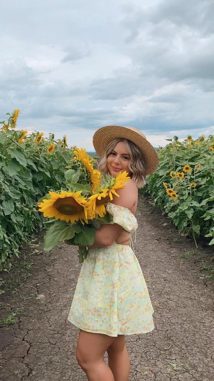 a woman in a sunflower dress and straw hat standing on a dirt road holding a bouquet of sunflowers