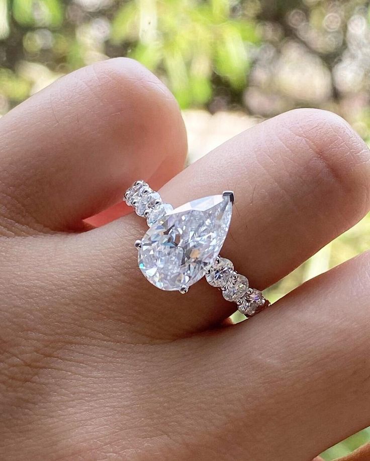 a woman's hand with a diamond ring on top of her finger, in front of some trees