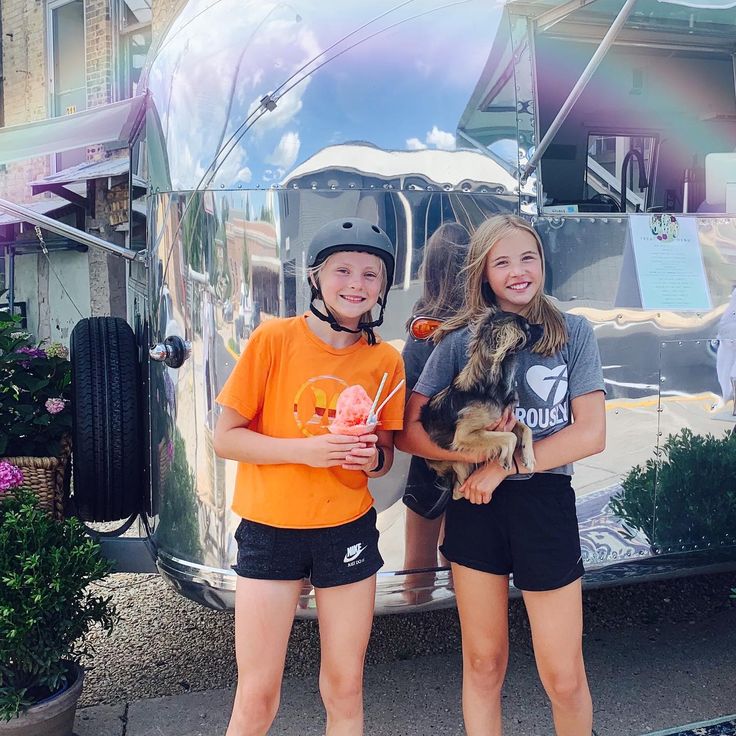 two girls standing next to each other in front of a silver trailer with an ice cream sundae on it