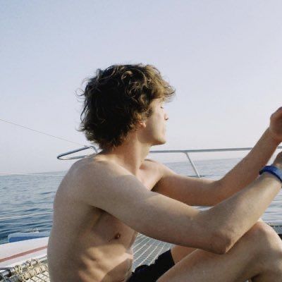 a shirtless man sitting on the bow of a boat holding a cell phone in his hand