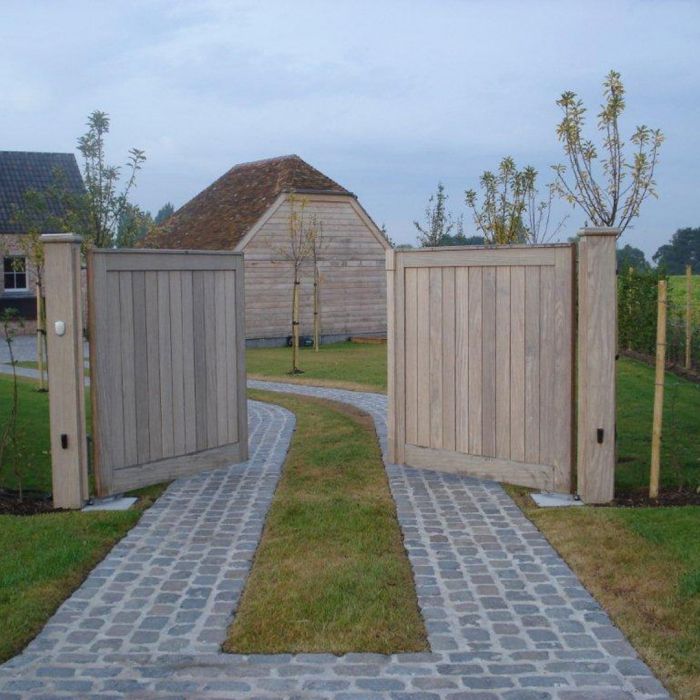 two wooden gates are open on a brick path leading to a barn and garden area