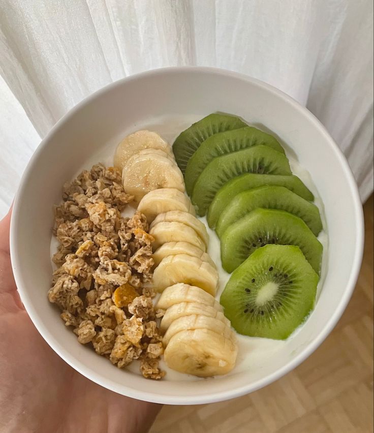 a person holding a bowl with cereal, kiwis and bananas in it on top of a wooden table