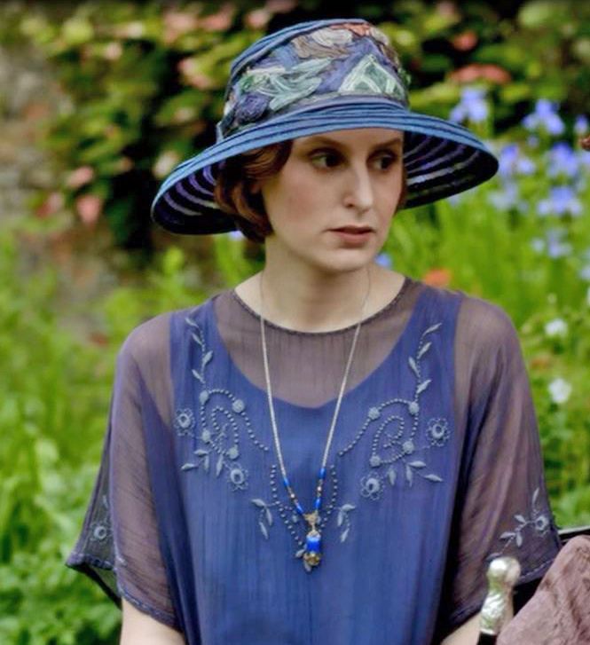 a woman wearing a blue hat and dress in front of green grass with flowers behind her