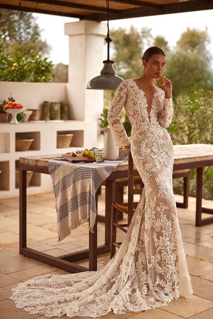 a woman standing in front of a table wearing a wedding dress with long sleeves and an open back