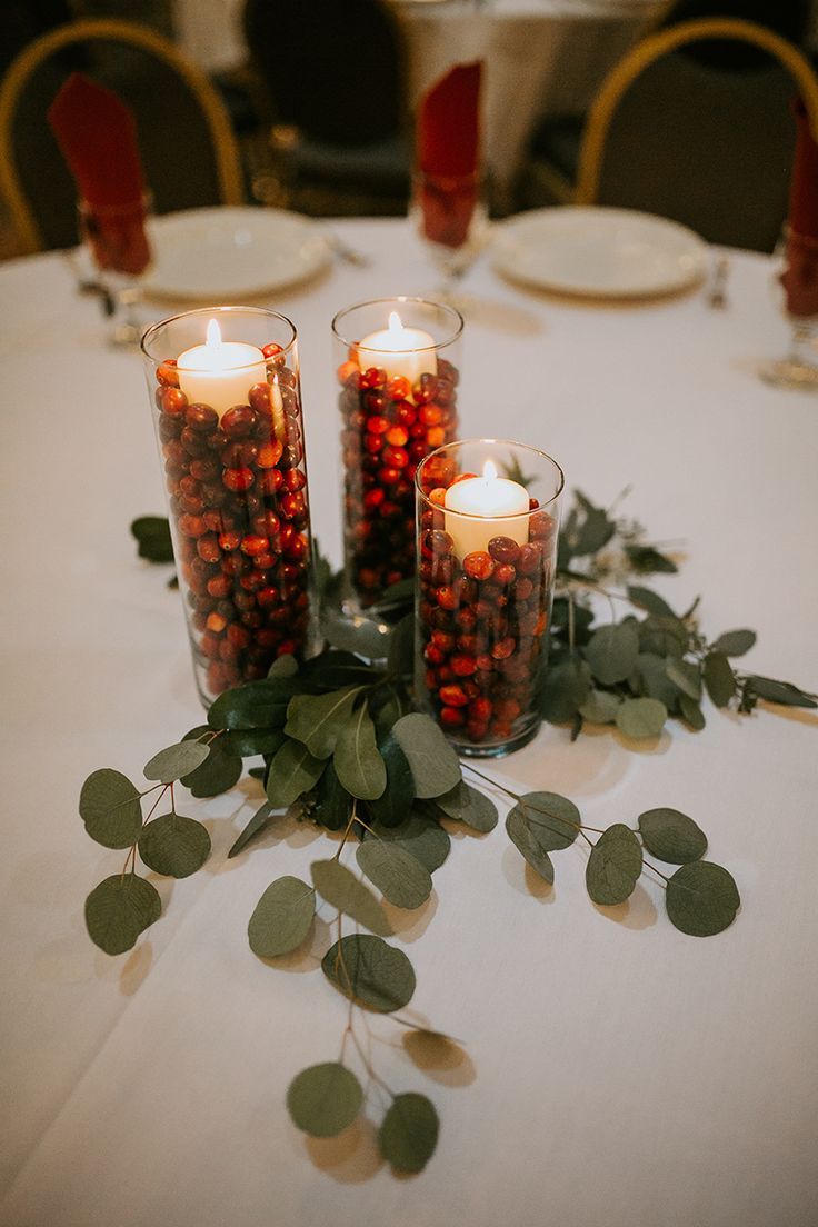 three candles are sitting on a table with greenery and red berries in glass vases