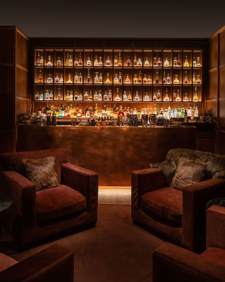 two brown chairs in front of a bar with bottles on the wall and shelves behind them