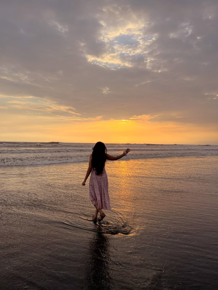 a woman standing on top of a sandy beach next to the ocean at sunset with her arms outstretched