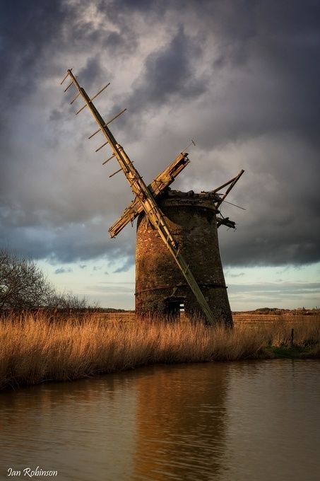 an old windmill sitting on top of a dry grass field next to a body of water