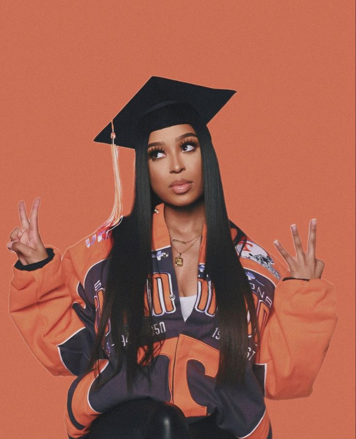 a woman with long black hair wearing an orange graduation gown and holding her hands up in the air