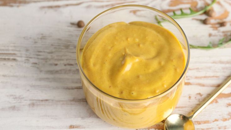 a glass filled with yellow liquid sitting on top of a wooden table next to a spoon