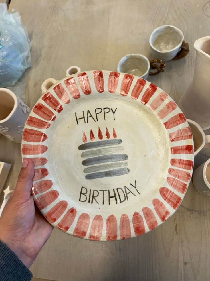 a person holding a plate with the words happy birthday written on it, surrounded by coffee cups and mugs