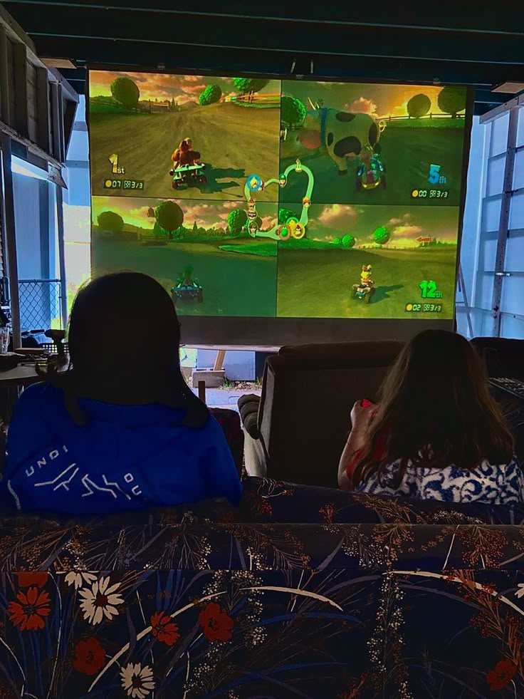 two people sitting in front of a large screen with mario kart racing on it