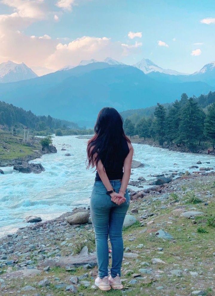 a woman standing on the side of a river with mountains in the backgroud