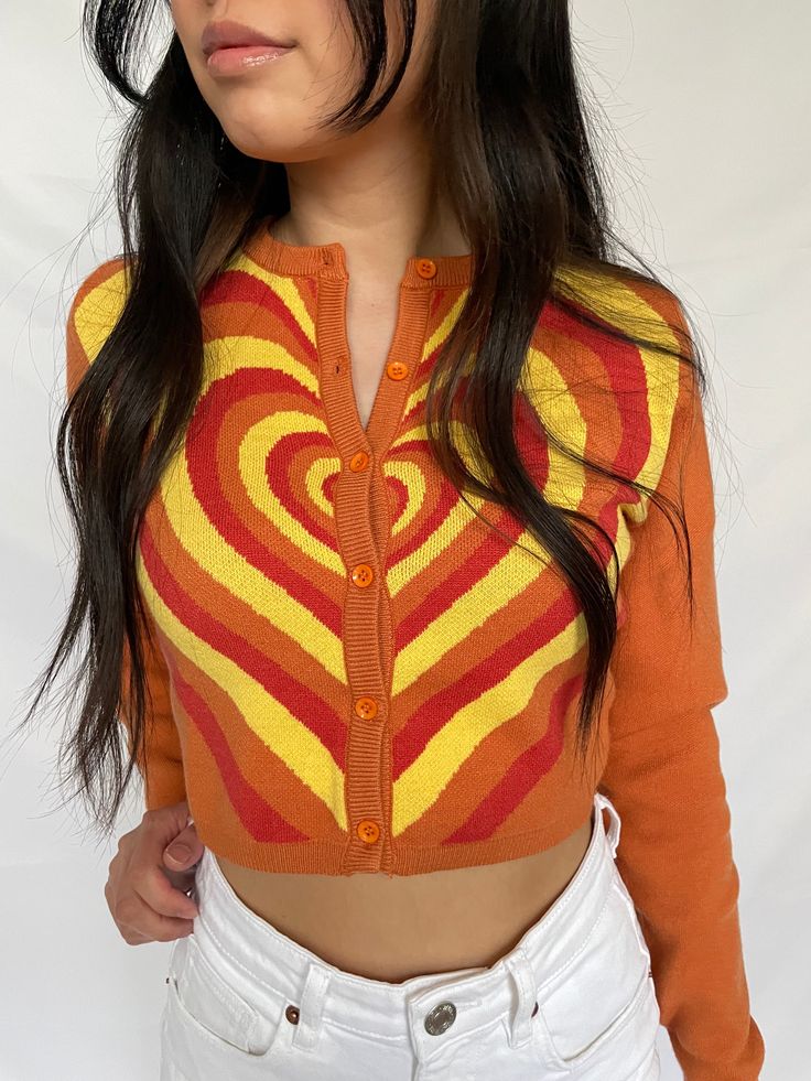 cropped orange cardigan with red, orange, and yellow heart design available in sizes xs, s, m, l model is 5'3" and wearing a size small Red Orange Outfit, Fitted Orange Cardigan For Spring, Fitted Heart Print Sweater For Fall, Retro Orange Cardigan For Winter, Trendy Fitted Orange Sweater, Fitted Trendy Orange Sweater, Orange Fitted Trendy Sweater, Retro Orange Spring Cardigan, Fitted Long Sleeve Orange Cardigan