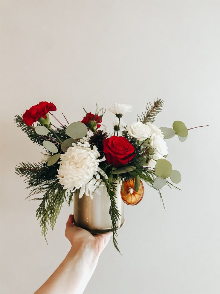 a person holding a vase with flowers and greenery in it on a white background