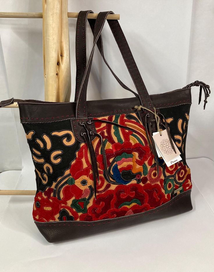 Pranee Brooklyn Abbey Tote Bag    Abbey, from Brooklyn Collection, is a boho-chic tote.  Its abstract hand-sewn applique crafted with dark brown leather gives it rarity, while its size lends to utility.  Pranee Handcrafted Unique Designer Bags are beautiful artisan-made bags completely handmade using genuine leather, hand sewn embroidery, and unique decorative stitching.  There is an incredible amount of care and attention to details taken in the craftsmanship of every Pranee bag.  Each piece is made with genuine leather, hemp or cotton, and unique hand-sewn embroidery and/or applique. Features * Zipper closure * Interior side pockets and zipper pocket * Genuine leather handle and accents  Specifications *approximate * Dimensions:  L 19" (top) 16" (bottom) x W 1" x H 14"  Materials * Hand- Hand-stitched Leather Tote Bags, Artisan Brown Shoulder Bag For Travel, Multicolor Leather Handle Bags For Fall, Multicolor Bags With Leather Handles For Fall, Fall Multicolor Bags With Leather Handles, Bohemian Satchel Bag For Fall, Traditional Brown Weekender Bag With Leather Handles, Bohemian Brown Weekender Bag With Leather Handles, Hand-stitched Leather Tote Satchel