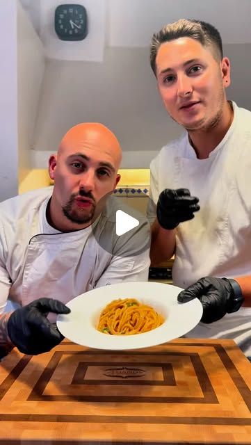 two men sitting at a table with a plate of spaghetti in front of them, and one man wearing black gloves