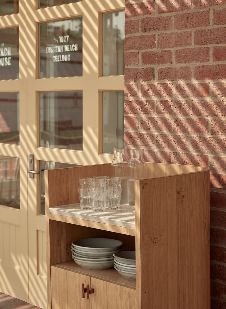 a wooden cabinet with plates and glasses on it in front of a brick wall that has glass windows