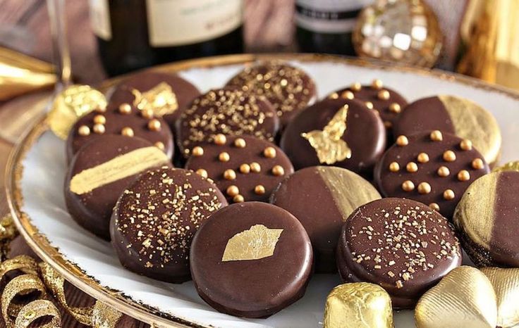 some chocolates are on a white plate with gold decorations and wine bottles in the background