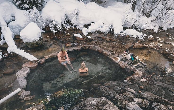 two people sitting on rocks in a hot tub surrounded by snow and ice covered trees