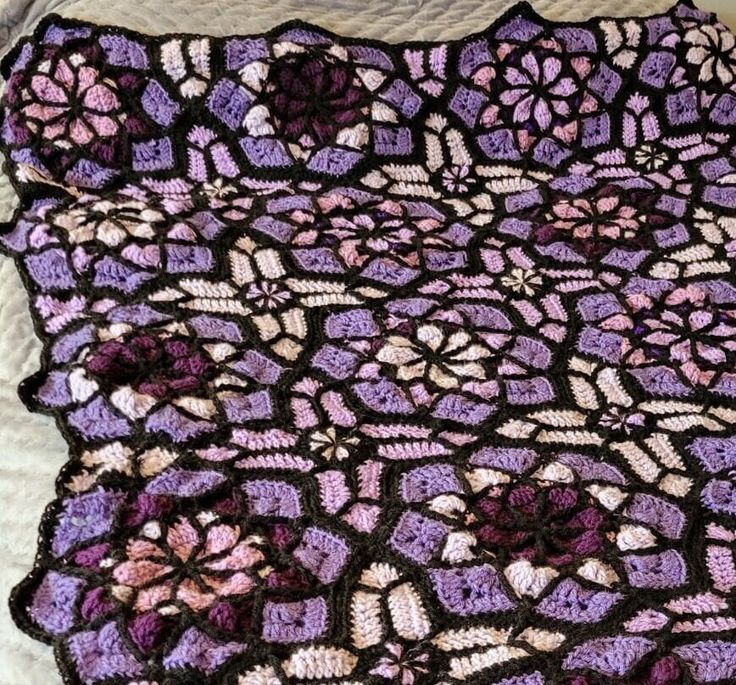 a crocheted blanket is laying on top of a white bed sheet with purple and pink flowers