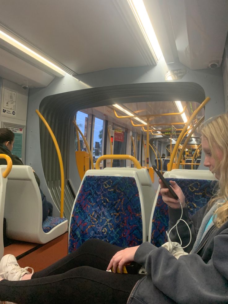 two people sitting on a bus looking at their cell phones and listening to headphones