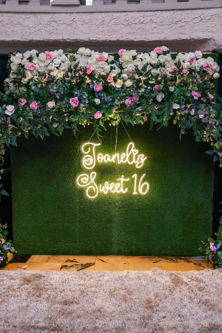 the neon sign is lit up in front of a green backdrop with pink and white flowers