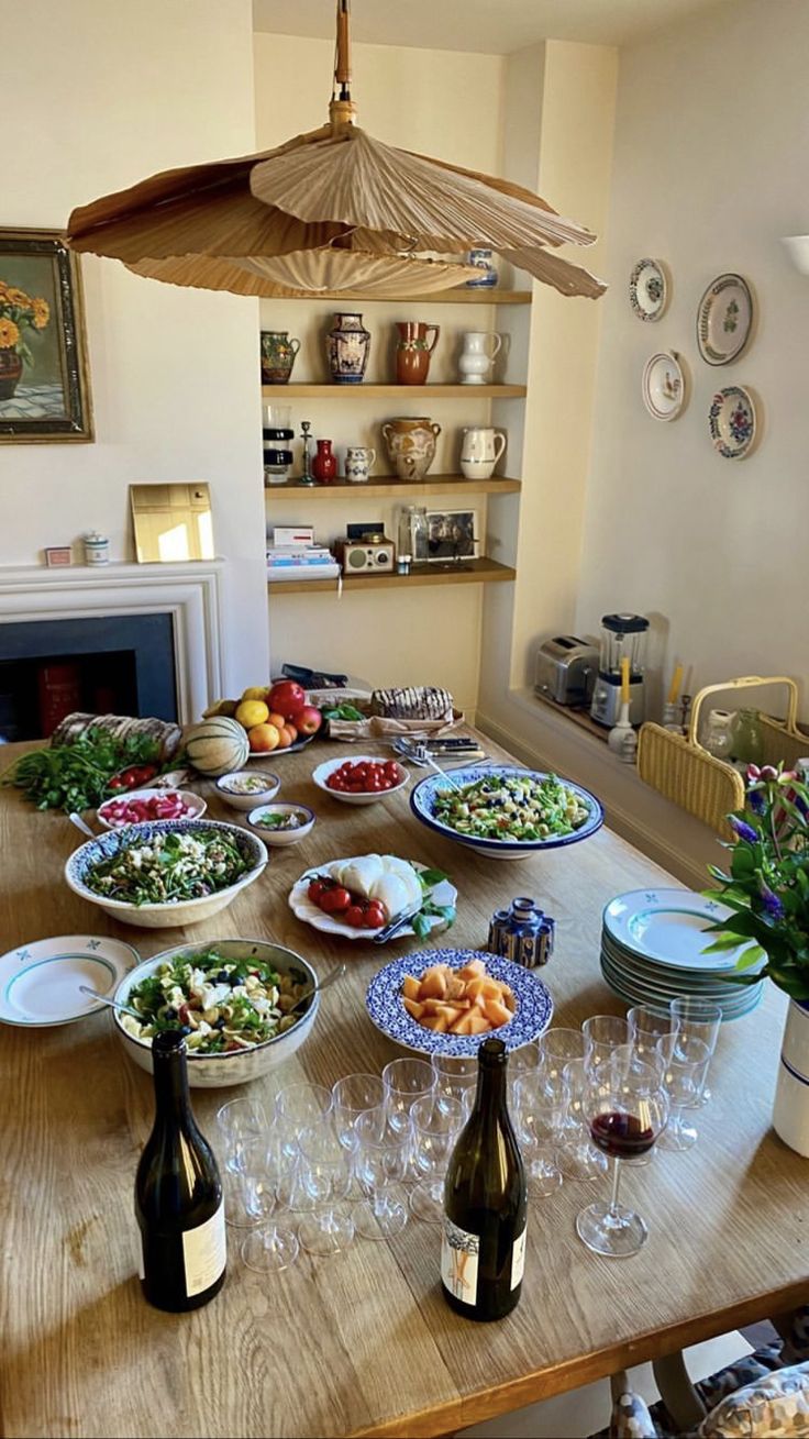 a wooden table topped with lots of plates and bowls filled with food next to bottles of wine