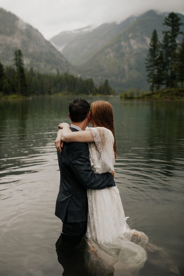 a man and woman are hugging in the water by some mountains with trees on it