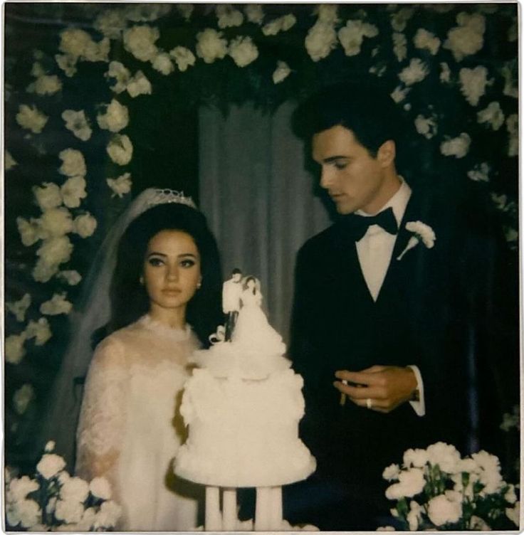 a man and woman standing next to each other in front of a white wedding cake