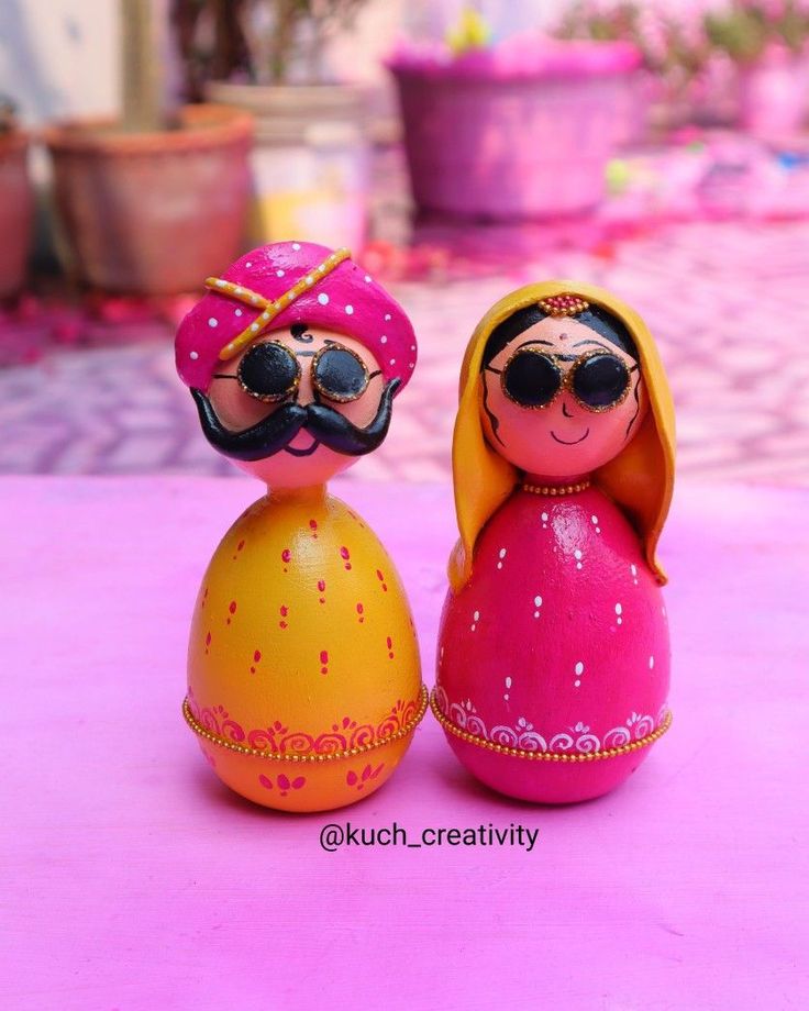 two wooden dolls sitting next to each other on a pink tableclothed surface with potted plants in the background