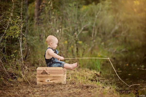 a baby sitting on top of a wooden box in the woods with a fishing rod