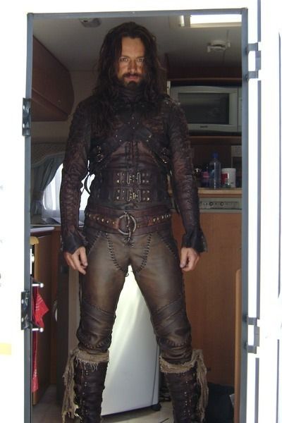 a man with long hair and beard standing in an open doorway wearing steampunk clothing