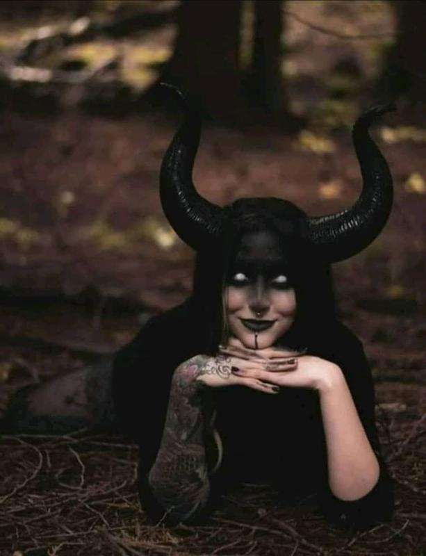 a woman with black makeup and horns on her face is laying in the woods while holding her hands to her chest