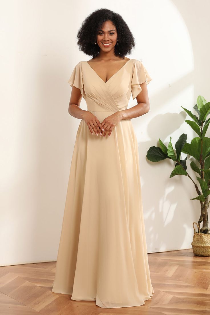 a woman in a tan dress standing on a wooden floor next to a potted plant