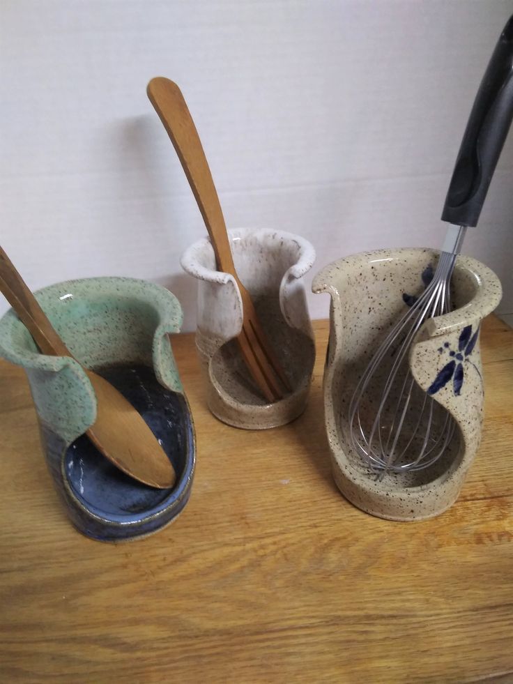 three ceramic utensils are lined up on a wooden counter top, one has a whisk in it and the other is empty