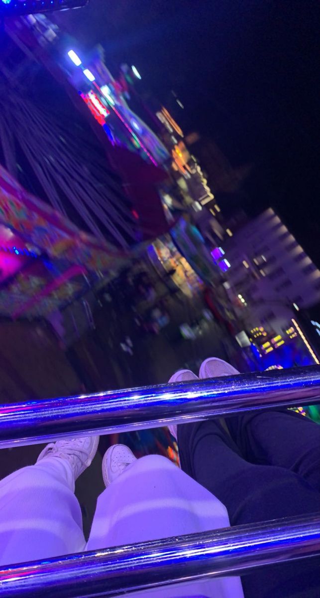 someone is sitting on the edge of a glass railing at night with lights in the background