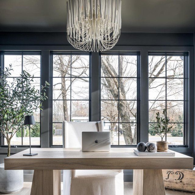 a desk with a laptop on it in front of two windows and a chandelier hanging from the ceiling