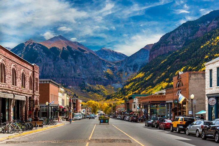 a street lined with parked cars in front of tall mountain range covered in fall foliage