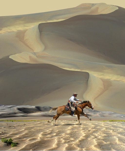 a man riding on the back of a brown horse across a sandy desert field with mountains in the background