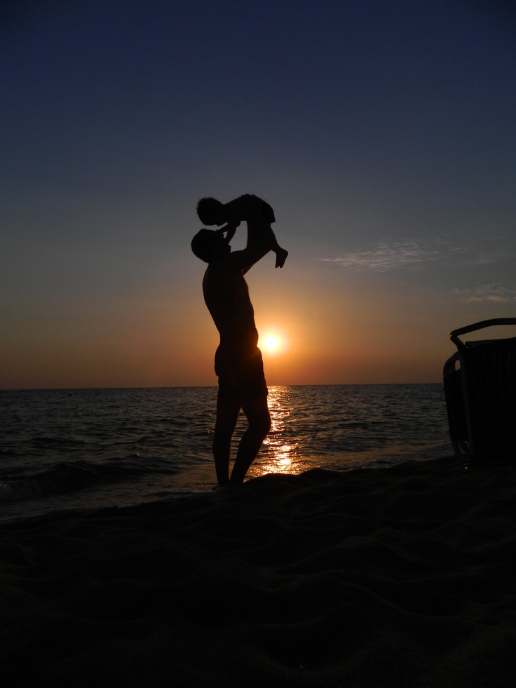 a man holding a child on his shoulders while standing in front of the ocean at sunset