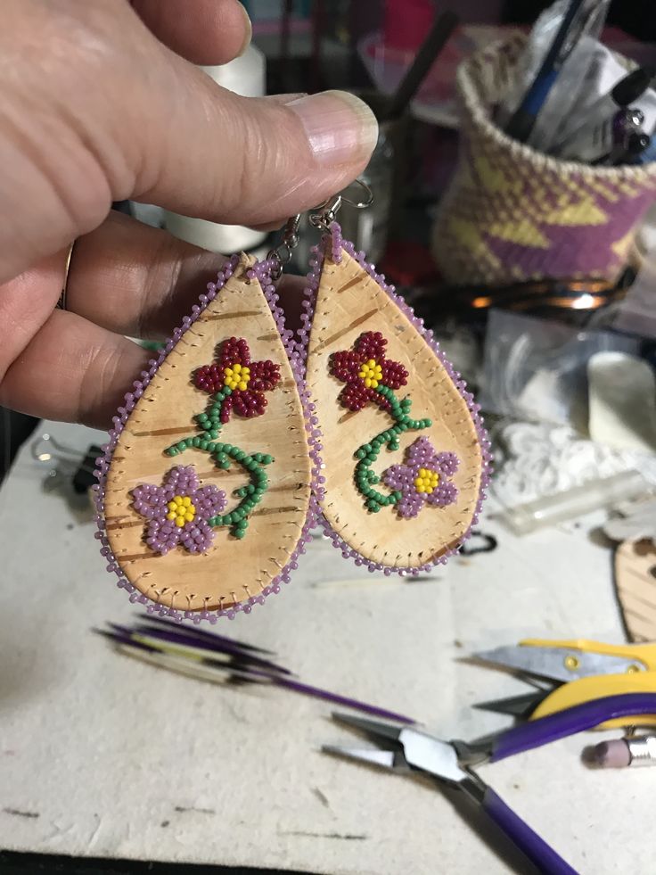 a person holding up two wooden earrings with beading on them and some scissors in the background