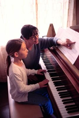 two people sitting at a piano with sheet music on the keyboard and one person standing next to it