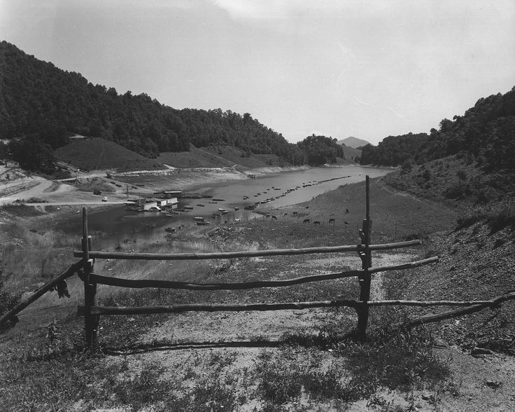 a black and white photo of a fence in front of a body of water surrounded by hills