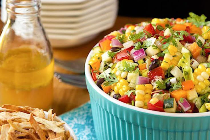 a bowl of corn salad next to a container of potato chips and a bottle of oil